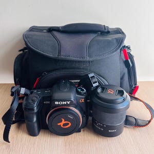 May include: A black Sony Alpha 200 digital SLR camera body with a lens cap, a separate black Sony 18-70mm lens with a cap, and a black camera bag with red accents. The camera features the Sony logo and Alpha branding on the front and lens.