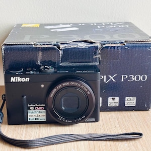 Op de afbeelding: Een zwarte Nikon Coolpix P300 digitale camera met polsband, voorzien van een Nikkor 4.2x groothoekzoomlens, CMOS-sensor en Full HD-filmfuncties. De camera staat voor de originele donkerblauwe productverpakking.