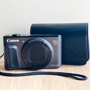 Może przedstawiać: Ciemnoszary aparat cyfrowy Canon PowerShot SX720 HS z obiektywem z 40-krotnym zoomem optycznym, teksturowanym uchwytem z przodu i czarnym paskiem na nadgarstek. Obok aparatu znajduje się pasujący ciemnoniebieski futerał z wytłoczonym napisem PowerShot.