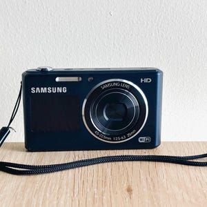 May include: A dark blue Samsung digital camera with a black wrist strap. The front features a Samsung lens with 4.5-22.5mm 1:2.5-6.3 25mm specifications, a silver shutter button, a flash, and white text reading Samsung, HD, and Wi-Fi.