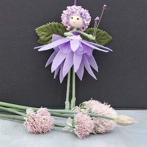 May include: A decorative doll resembling a flower fairy with a light purple petal skirt, a textured lavender headpiece, and green leaf-shaped wings. The figure has thin green limbs and holds a small stem with purple buds, positioned above three light pink chive blossoms.