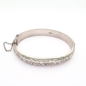 May include: A silver bangle bracelet with an engraved floral pattern. The hinged bracelet has a safety chain and a clasp closure. The design features detailed leaf and floral motifs. The bracelet is displayed on a white background.