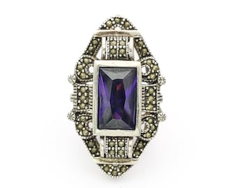 May include: A silver-toned ring with a rectangular, deep purple gemstone at its center. The ring features an ornate design with small, round, silver-colored accents and a decorative frame around the gemstone. The ring is set against a white background.