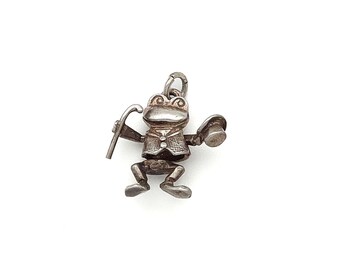 May include: A silver-toned metal charm shaped like a frog wearing a vest and holding a cane in one hand and a top hat in the other. The frog has a textured surface, large eyes, and a small jump ring attached to the top for hanging as jewelry.