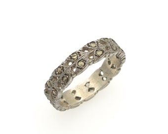May include: A silver-toned band ring featuring a repeating openwork leaf pattern set with small, dark, faceted stones. The metal surface has a textured, matte finish, and the design extends around the entire circumference of the circular ring.