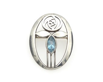 May include: An oval silver brooch featuring a stylized rose cutout at the top, two horizontal leaves, and a central marquise-cut light blue gemstone. The metal frame is smooth and polished, enclosing the floral design and the blue stone.