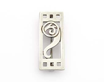May include: A rectangular silver-toned brooch with an abstract design. The brooch features a stylized swirl within a rectangular frame, with small square cutouts along the edges. The pin is a vintage accessory.