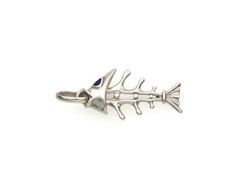 May include: A silver-toned metal pendant shaped like a fish skeleton. The design features a small, dark blue inlay for the eye and a circular loop at the head for attachment to a chain. The structure shows the spine and ribs of the fish.