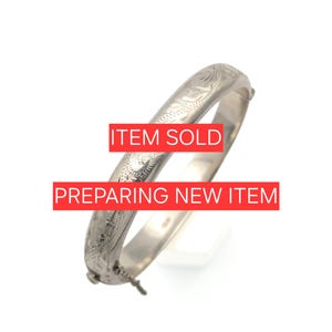 Könnte beinhalten: Ein silberfarbener Klapparmreif mit einem komplizierten gravierten Blumenmuster auf der Außenfläche. Das Armband verfügt über einen sicheren Verschlussmechanismus und zeigt den Text "ITEM SOLD" und "PREPARING NEW ITEM" in Weiß.