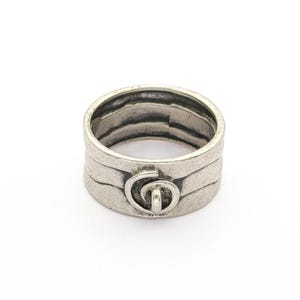 May include: A silver ring with a textured, layered design. The ring features a central decorative element with a circular motif. The ring's band has a slightly worn appearance, suggesting an antique or vintage style. The ring is set against a white background.