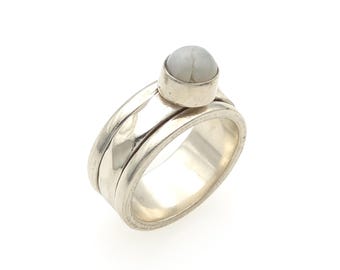May include: A silver ring featuring a wide, polished band with a smaller, thinner spinning band layered on top. A single, round, light gray stone with subtle white veining is set in a smooth silver bezel atop the spinning band.