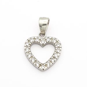 Puede incluir: Colgante plateado en forma de corazón con un lazo en la parte superior. El corazón está delineado con pequeñas piedras preciosas transparentes. El colgante se muestra sobre un fondo blanco.