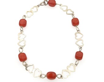 May include: A bracelet featuring a repeating pattern of silver-toned metal heart-shaped links alternating with smooth, rounded, deep orange-red beads. The jewelry piece is secured with a small circular metal clasp at the top of the circular arrangement.