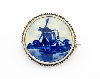 May include: A round brooch with a blue and white windmill design. The image is set in a silver-toned metal frame with a twisted rope-like border. The brooch has a pin on the back for attachment.