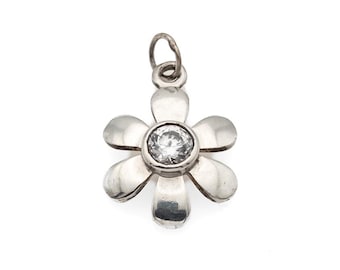 May include: A silver-toned metal flower pendant featuring six smooth, rounded petals arranged around a central, clear, round-cut faceted gemstone. The pendant includes a small circular jump ring at the top for attachment to a chain or cord.