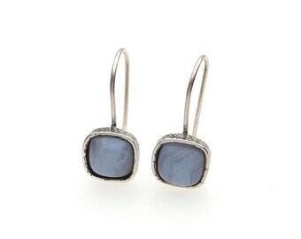 May include: A pair of drop earrings featuring square-shaped, light blue stones set in textured, dark silver-toned metal frames. Each earring has a curved wire hook for attachment, showcasing a smooth, polished surface on the stones.
