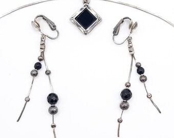 May include: A silver-toned jewelry set featuring a pendant necklace and clip-on earrings. The pendant has a black square stone set in a diamond-shaped frame. The earrings and pendant have black and silver beads.
