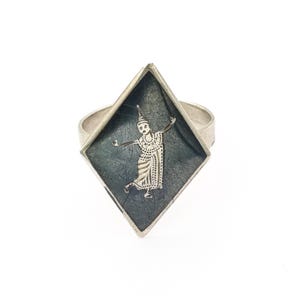 Könnte beinhalten: Silberring mit rautenförmiger Fassung und dunklem Hintergrund. Eine detaillierte Silberfigur einer Tänzerin ist auf der Fassung zentriert. Der Ring hat einen breiten Ring und eine Vintage-Ästhetik.