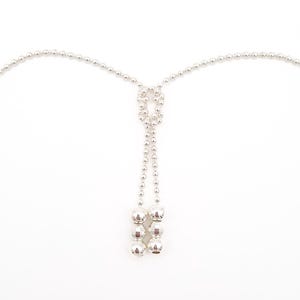 May include: A silver-colored body jewelry piece, likely a necklace or body chain, against a white background. The design features small, round beads connected by a delicate chain, with a cluster of larger, faceted beads at the bottom.