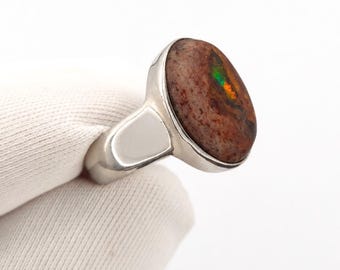 May include: A silver ring featuring a large, oval-shaped boulder opal gemstone. The stone displays a mottled brown and tan surface with a vibrant, iridescent green and orange flash of color embedded within the center of the natural stone texture.