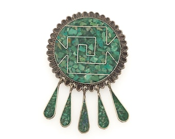 May include: A circular silver brooch featuring a central inlay of crushed green malachite stone arranged in a geometric spiral pattern. The outer edge has a scalloped border, and five teardrop-shaped silver dangles, also inlaid with green malachite, hang from the bottom of the main circular piece.