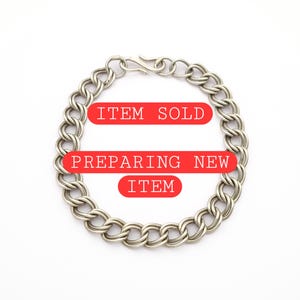 Pode incluir: Uma pulseira de corrente de metal prateado com um design de elos duplos e um fecho de gancho. A imagem inclui sobreposições retangulares vermelhas com texto branco que diz ITEM SOLD e PREPARING NEW ITEM sobre o centro da pulseira.