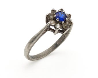 May include: A silver-toned ring featuring a central round blue stone surrounded by a cluster of smaller, clear, circular stones. The metal band has a weathered, matte finish, and the setting is elevated above the band in a floral-like arrangement.