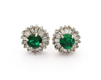 May include: A pair of stud earrings featuring a central, round, emerald-green gemstone surrounded by a halo of smaller, baguette-cut, clear gemstones. The earrings have a silver-colored metal setting and are displayed against a white background.