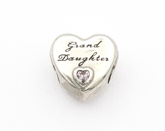 May include: A silver heart-shaped charm with the words "Grand Daughter" engraved on the front. A small pink heart-shaped gem is set in the center. The charm is designed to be a pendant or bead.