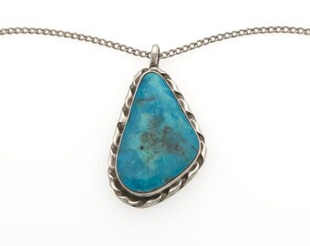 May include: A silver pendant necklace featuring an irregular, teardrop-shaped turquoise stone with dark matrix inclusions. The stone is set in a polished silver bezel with a twisted rope-style border, suspended from a delicate silver chain.
