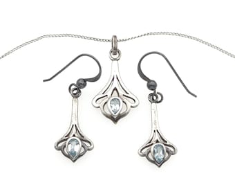 May include: A jewelry set consisting of a silver pendant on a thin chain and two matching drop earrings. Each piece features an openwork metal design with a single, small, light blue, pear-shaped gemstone set at the base of the silver frame.