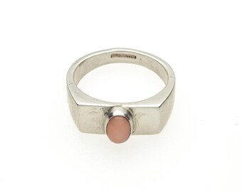 May include: A silver ring featuring a smooth, oval-shaped, light pink stone set in the center of a wide, polished metal band. The interior of the band displays small, engraved hallmarks indicating the metal purity and origin of the piece.