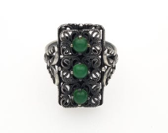 May include: A silver-toned ring featuring an elongated rectangular face with intricate filigree metalwork. Three smooth, round, vibrant green cabochon stones are set vertically in a line along the center of the detailed, dark-oxidized metal frame.
