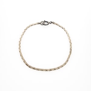 Puede incluir: Una pulsera de cadena de espiga en tono plateado con un diseño de eslabones planos y tejidos con un acabado metálico pulido. La pulsera se asegura con un pequeño cierre de resorte circular en la parte superior, creando un bucle flexible.
