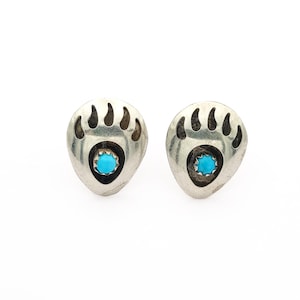 Könnte beinhalten: Ein Paar silberfarbene Ohrstecker in Form von Bärenpfoten. Jeder Ohrring hat ein Bärenpfoten-Design mit einem türkis farbenen Stein in der Mitte. Die Ohrringe sind vor einem weißen Hintergrund platziert.