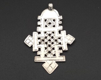 May include: A silver-toned metal pendant shaped like an intricate Ethiopian cross. The design features a central lattice pattern, geometric cutouts, and a small loop at the top for suspension. The surface has a metallic finish with engraved details.
