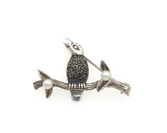 May include: A silver-toned brooch shaped like a bird perched on a textured branch. The bird's body is encrusted with small dark gray stones, and two round white pearls are set on the branch, one on each side of the bird, with a pin back.