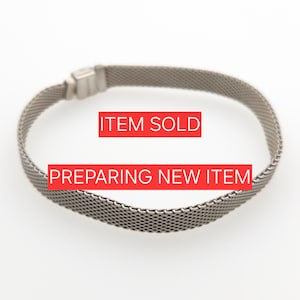 Op de afbeelding: Een zilverkleurige metalen mesh armband met een rechthoekige sluiting. De afbeelding bevat rode rechthoekige overlays met witte tekst die ITEM SOLD en PREPARING NEW ITEM leest, geplaatst over het midden van de armband.