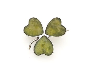 May include: A silver brooch shaped like a three-leaf clover, featuring three heart-shaped segments filled with mottled, translucent olive-green stone inlays. The metal frame outlines each leaf, and a small curved stem extends from the bottom left.
