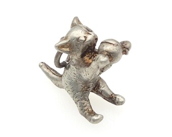 May include: A silver-toned metal charm shaped like a small cat sitting upright. The cat is depicted with its front paws raised, holding a small round ball. The surface of the charm features textured detailing to represent fur, with a small loop for attachment.