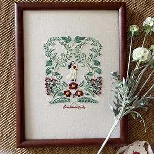 Puede incluir: Un bordado enmarcado que representa a una pareja de pie en un arreglo floral en forma de corazón. El bordado está sobre una tela de lino blanco y el marco es de madera marrón. El texto "Love never fails" está bordado debajo de la pareja.