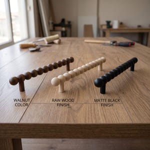 May include: Three linear cabinet pulls with a spherical bead design. The left pull is walnut color, the center pull is raw wood finish, and the right pull is matte black finish. Each handle features two mounting posts for attachment to furniture.