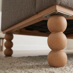 May include: Close-up of a furniture piece with a light brown wooden frame and legs. The legs are a stacked ball design. The upholstery is a neutral grey fabric. The piece is sitting on a light-coloured rug.