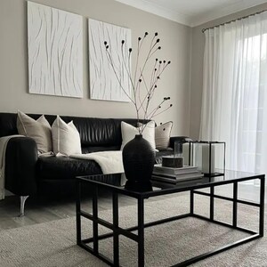 Peut inclure: Salon moderne avec un canapé en cuir noir orné de coussins et d'un plaid. Une table basse noire avec un plateau en verre repose sur un tapis gris clair. Deux œuvres d'art murales blanches abstraites et un vase noir avec des branches complètent le décor.