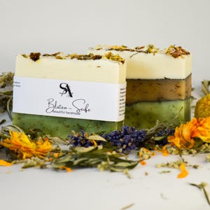 Könnte beinhalten: Zwei handgemachte Seifen mit Schichten in Grün, Braun und Creme, verziert mit getrockneten Blumen. Die Seifen haben ein weißes Etikett mit dem Text "Blüten-Seife". Die Seifen sind von getrockneten Kräutern und Blumen umgeben.