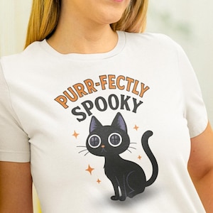 May include: White t-shirt with a cartoon black cat illustration and the text "Purr-fectly Spooky" in orange and black. The cat has button eyes and is surrounded by orange stars. The shirt is paired with blue jeans.