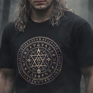 May include: Black t-shirt featuring a gold circular design with geometric shapes, symbols, and a skull. The person in the image has long, silver hair and is looking directly at the viewer. The background is a blurred forest.