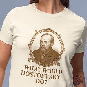 May include: Ivory-colored t-shirt featuring a sepia-toned portrait of Fyodor Dostoevsky within an ornate frame. Below the portrait, the text reads "WHAT WOULD DOSTOEVSKY DO?" The shirt is a classic crew neck style.