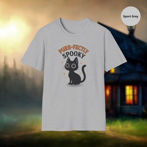 Może przedstawiać: Szara koszulka sportowa z ilustracją czarnego kota i napisem "Purr-fectly Spooky" w kolorze pomarańczowym. Projekt zawiera pomarańczowe akcenty w postaci gwiazd. Koszulka ma okrągły dekolt.