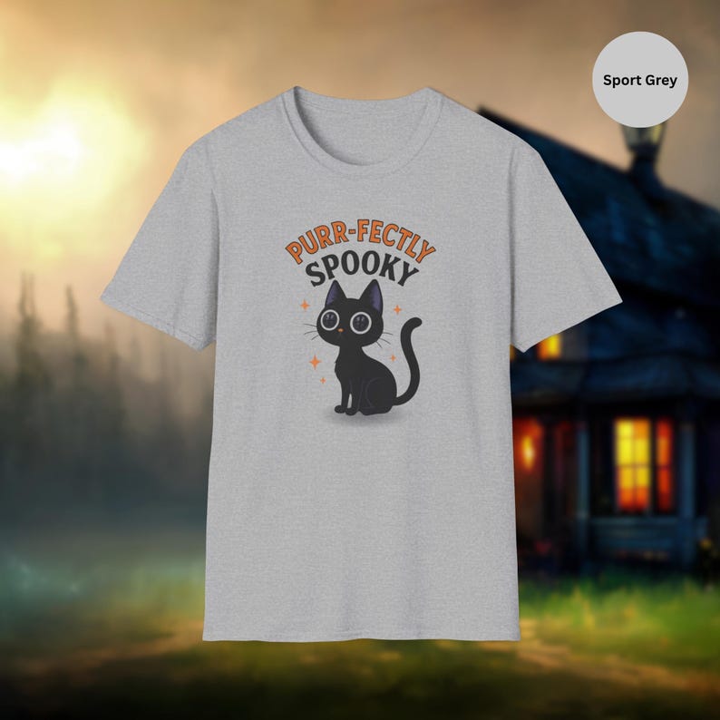 Może przedstawiać: Szara koszulka sportowa z ilustracją czarnego kota i napisem "Purr-fectly Spooky" w kolorze pomarańczowym. Projekt zawiera pomarańczowe akcenty w postaci gwiazd. Koszulka ma okrągły dekolt.